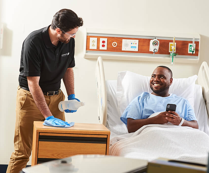 An Aramark employee disinfects the surfaces of a hospital room next to a patient lying in the bed.